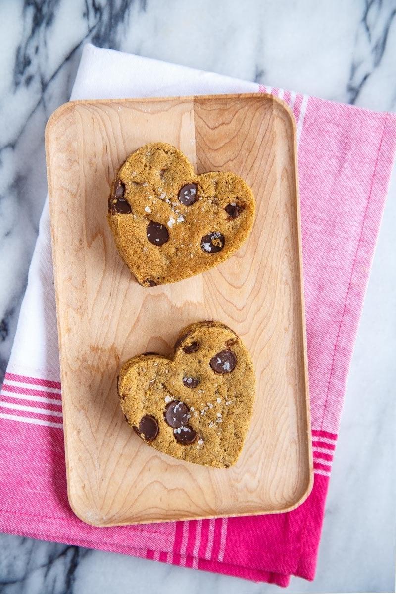 Due biscotti con gocce di cioccolato ritagliati a forma di cuore su un vassoio di legno.