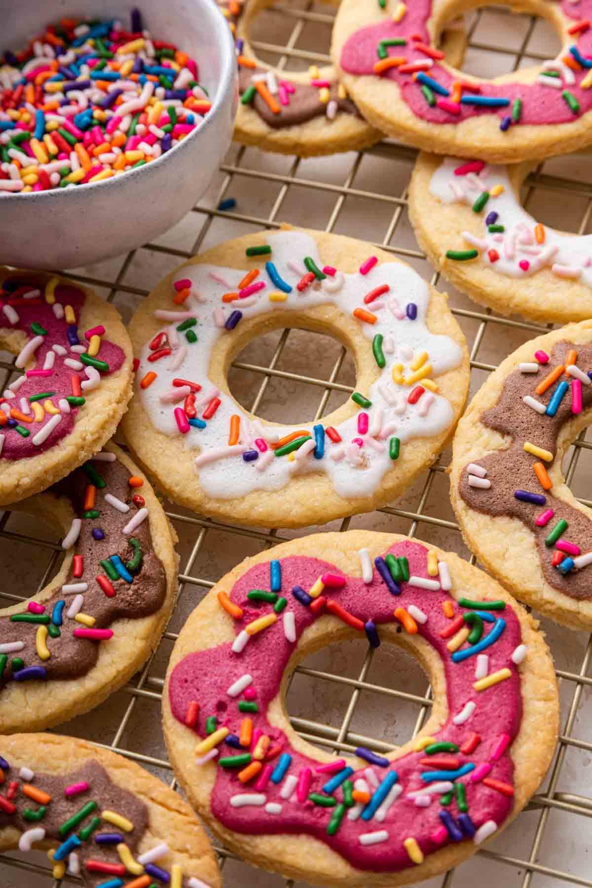 Biscotti Ciambella 2 Donut cookies with pink, white and brown frosting and sprinkles on a wire rack.