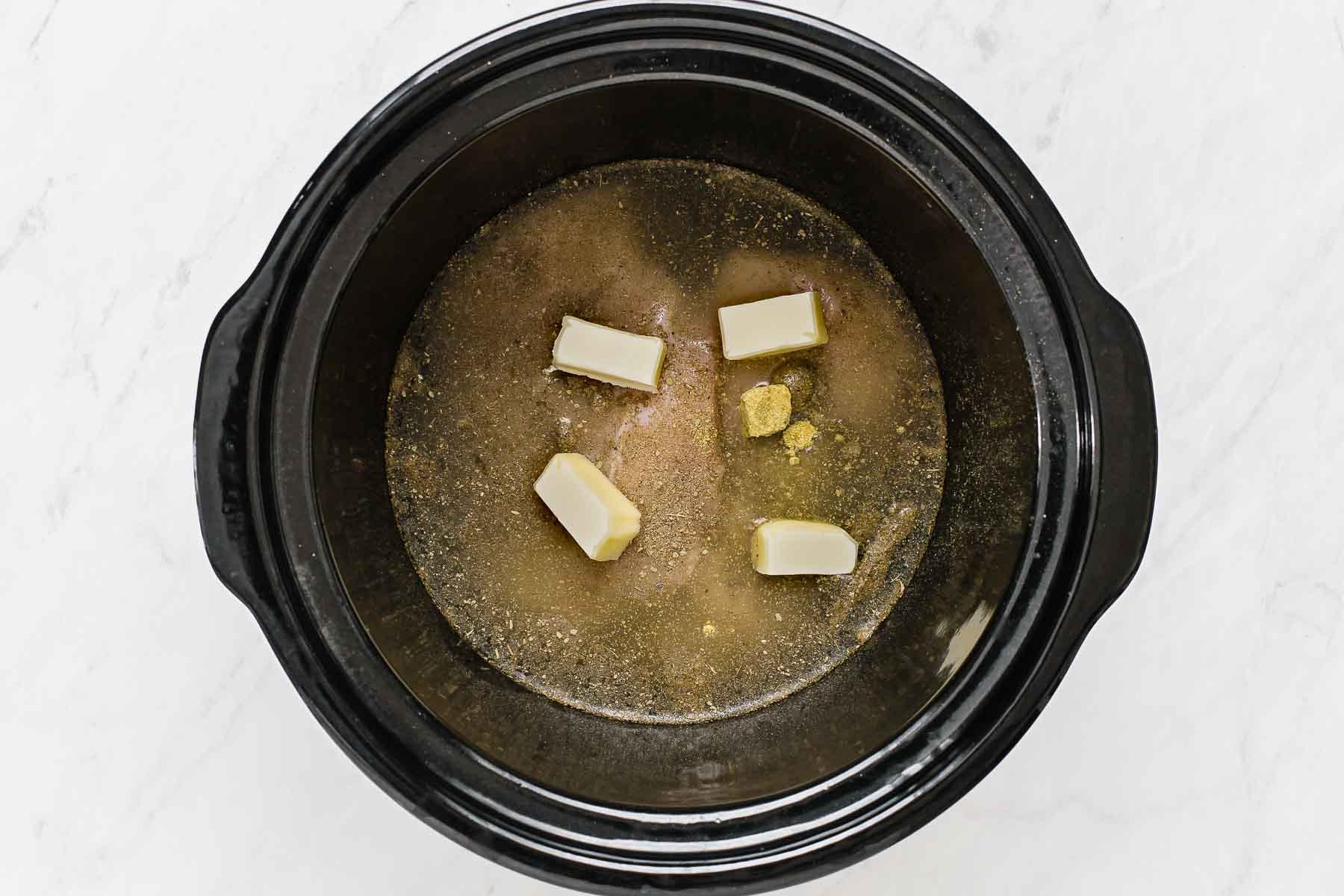 Pollo Cremoso in Pentola a Cottura Lenta 12 Petti di pollo con liquido e fette di burro sopra in slow cooker.