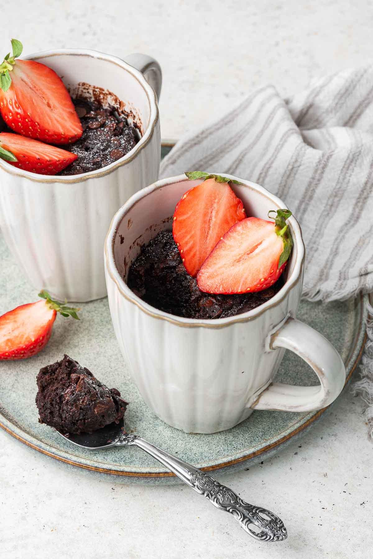 Ricetta Torta al Cioccolato in Tazza 3 Due tazze da caffè con un dessert scuro guarnito con fragole e un cucchiaino a lato.
