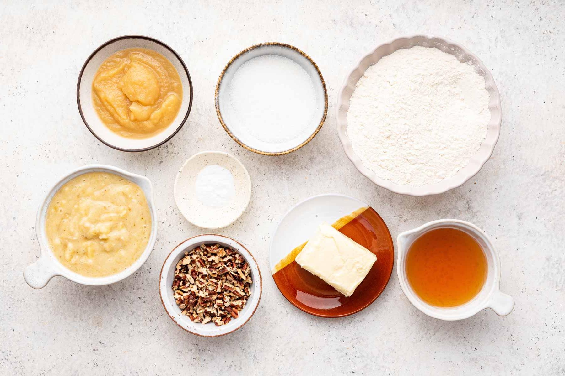 Pane alle banane senza uova 4 Ciotole bianche di ingredienti per la cottura su un bancone bianco.
