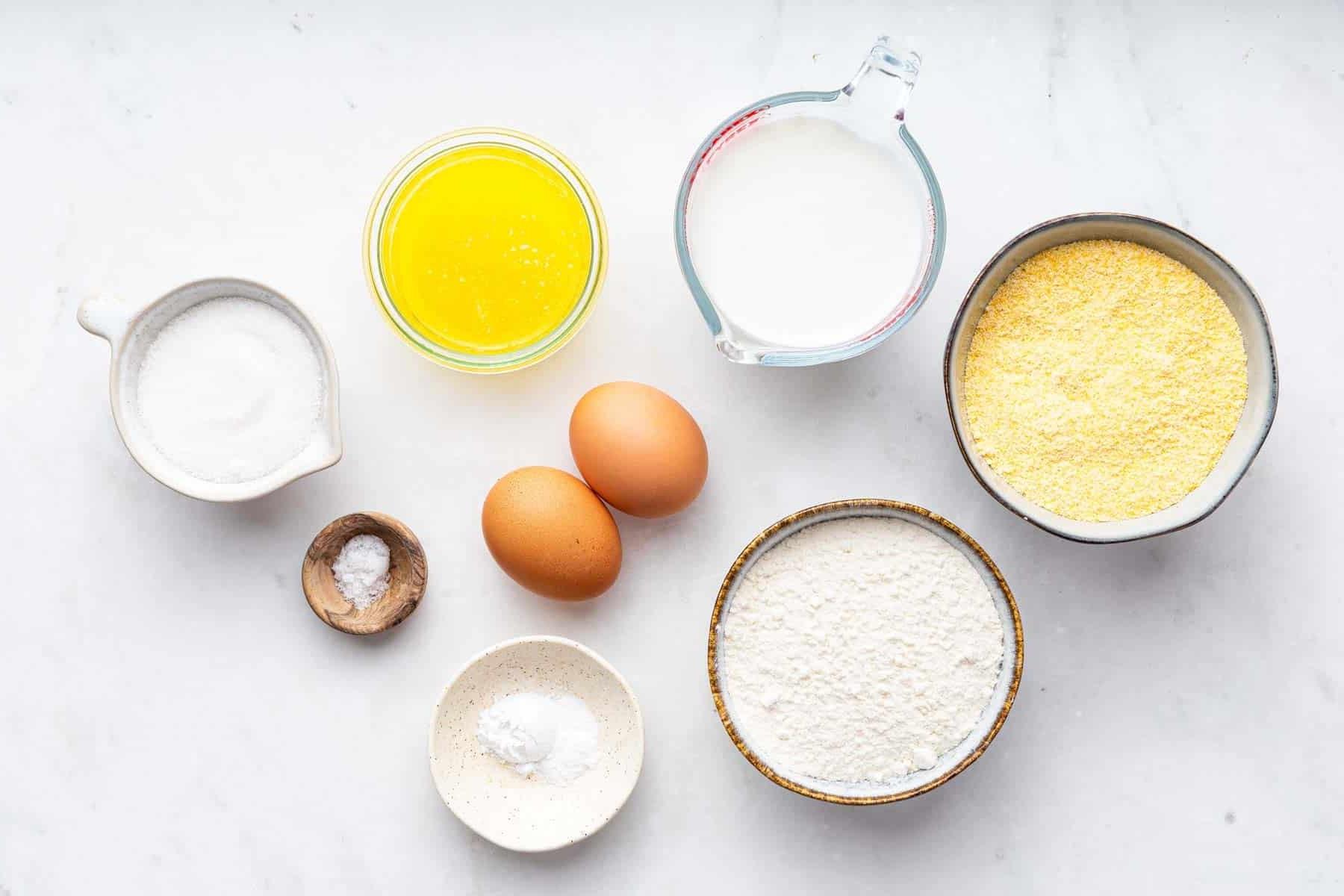 Ricetta Muffin al Mais 4 Ciotole di ingredienti bianchi e due uova sul bancone della cucina.