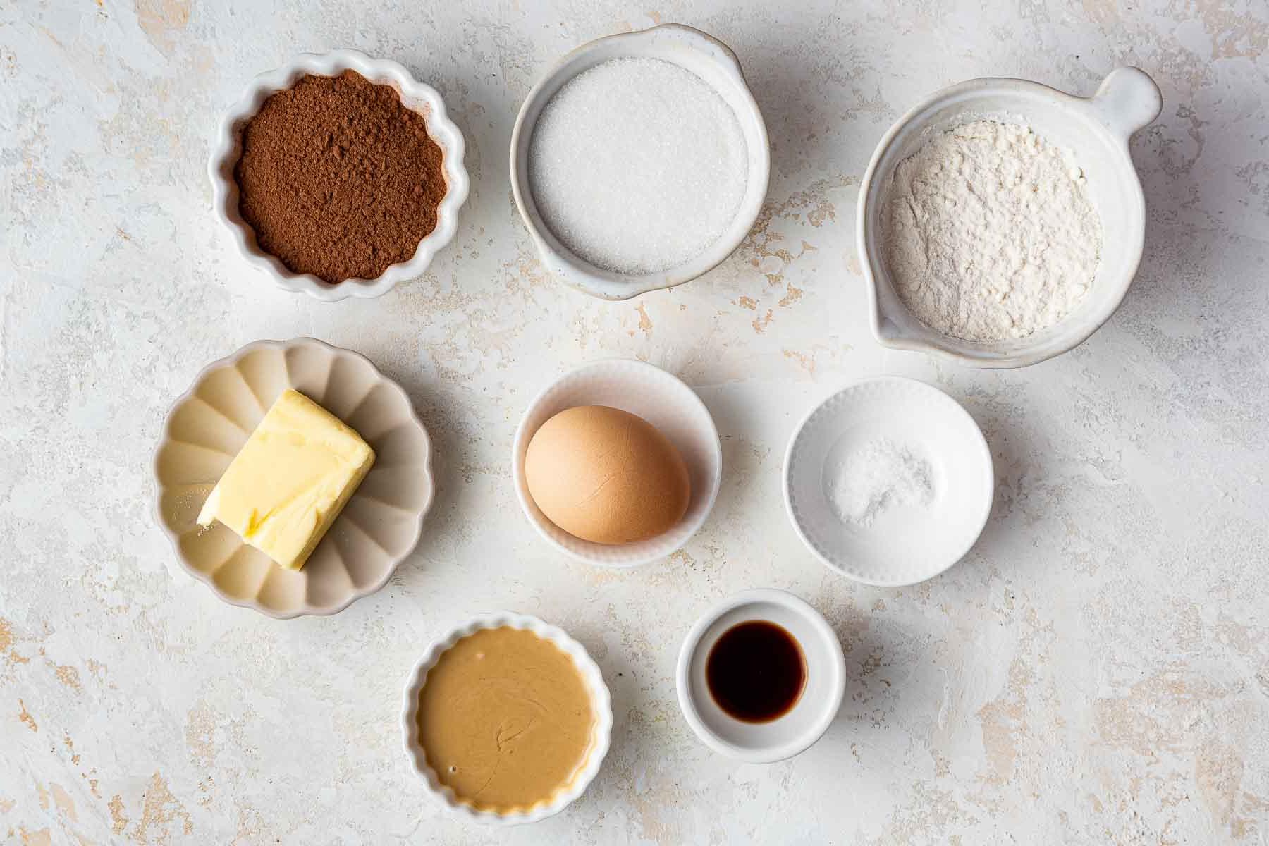 Ciotole di farina, zucchero, cacao in polvere, tahini e burro su superficie di marmo.