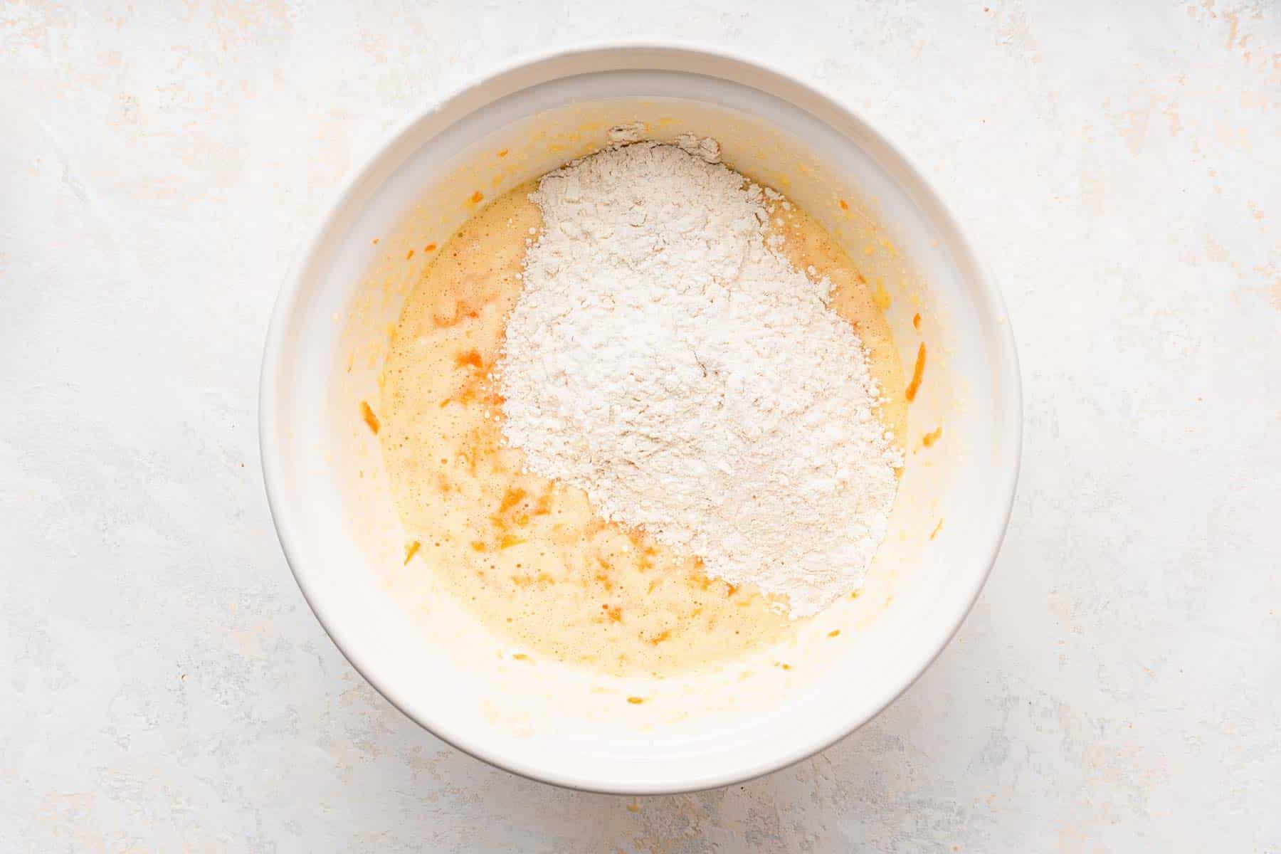 **Tortina di Carote con Glassa al Formaggio Cremoso al Caramello** 6 Ingredienti secchi delicatamente cosparsi sopra gli ingredienti umidi in una ciotola bianca, pronti per essere amalgamati.