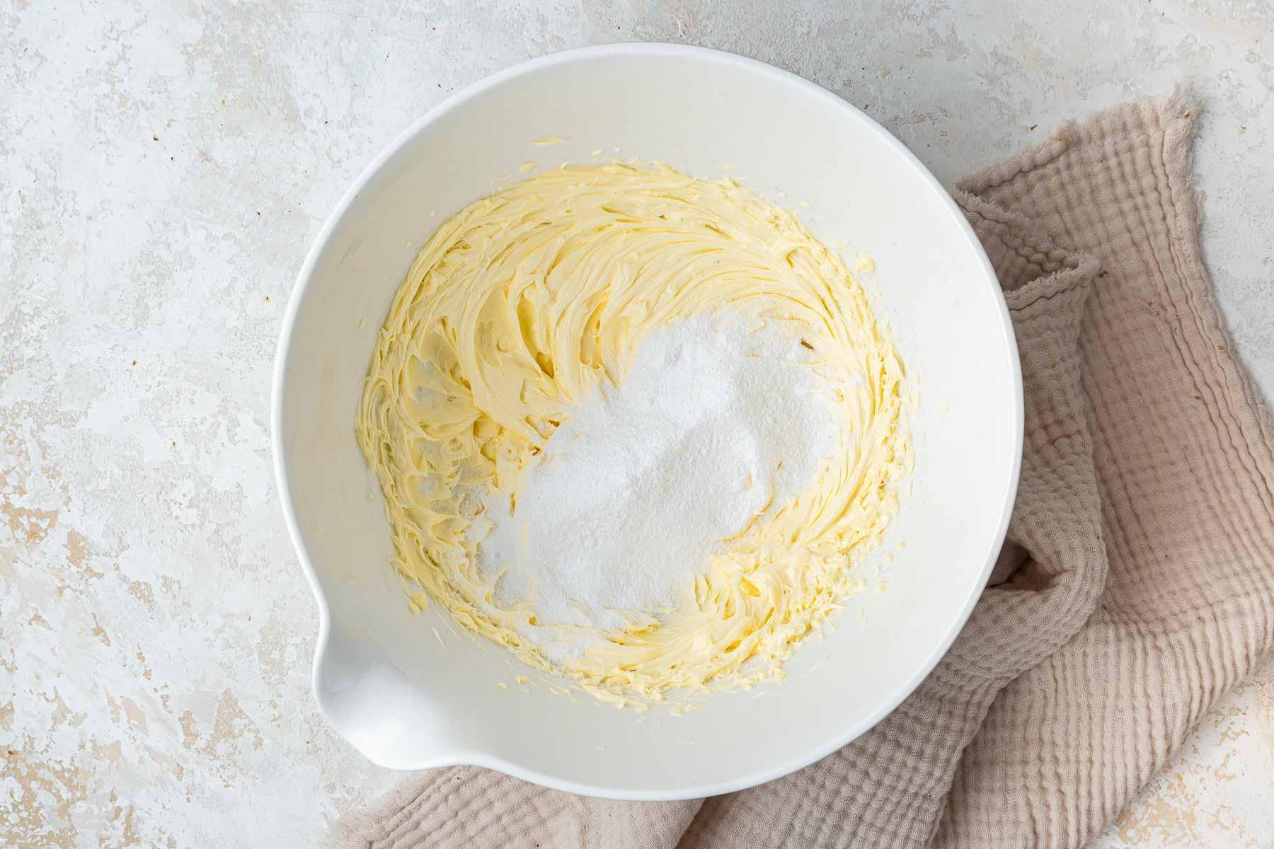Powdered sugar added to a bowl of beaten butter in white mixing bowl.