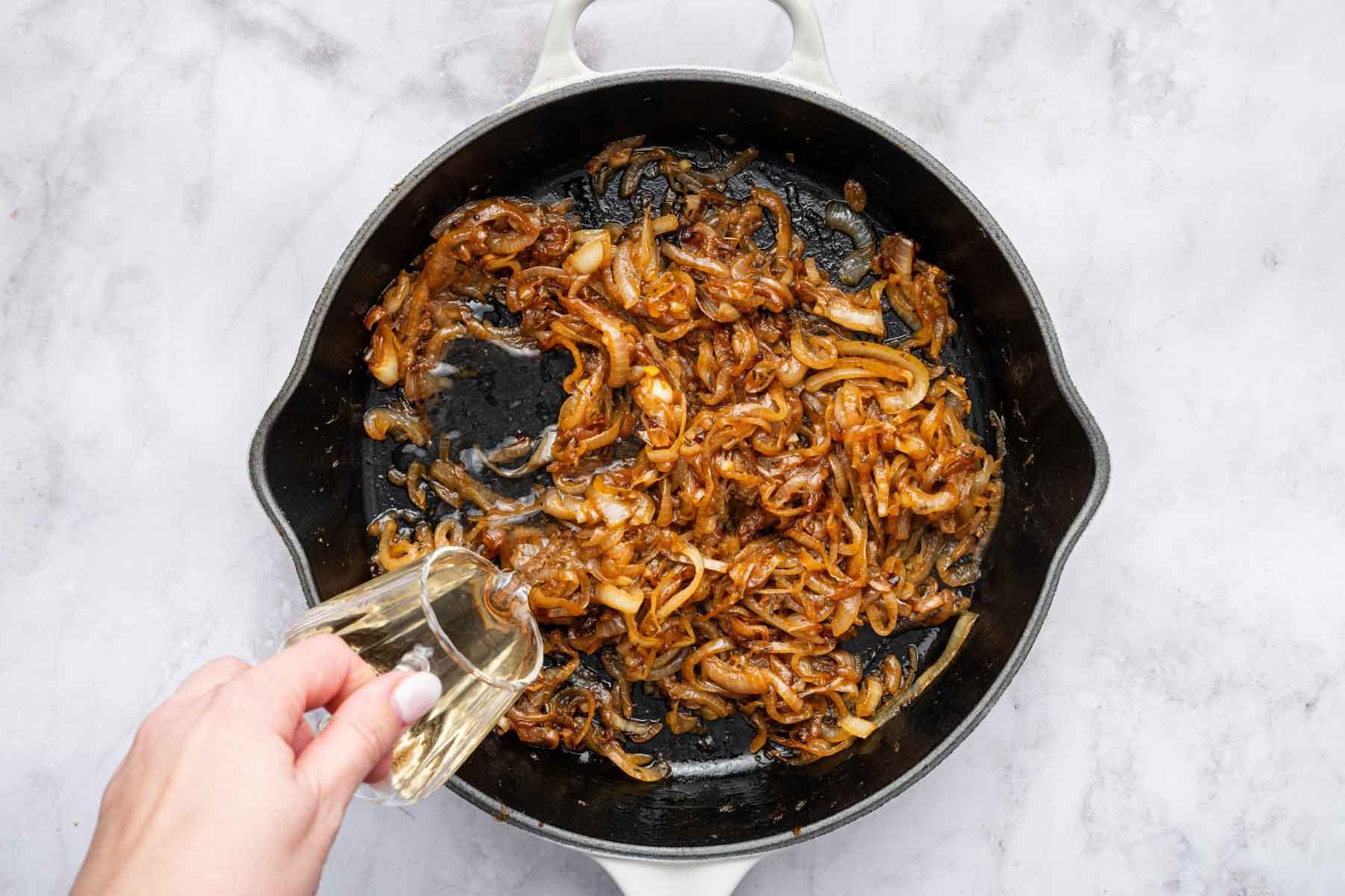 Ricetta del Pollo alla Cipolla Francese 6 Mano che versa vino bianco in una padella di ghisa con verdure cotte.