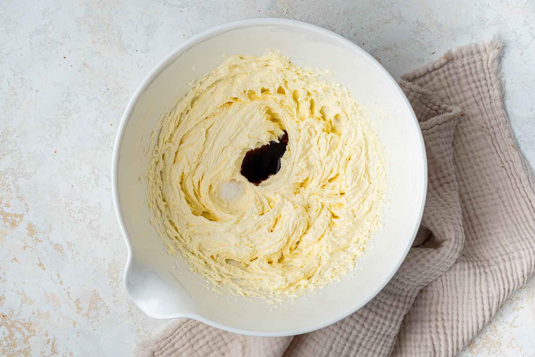 Drops of vanilla and salt in a yellow frosting in white mixing bowl.