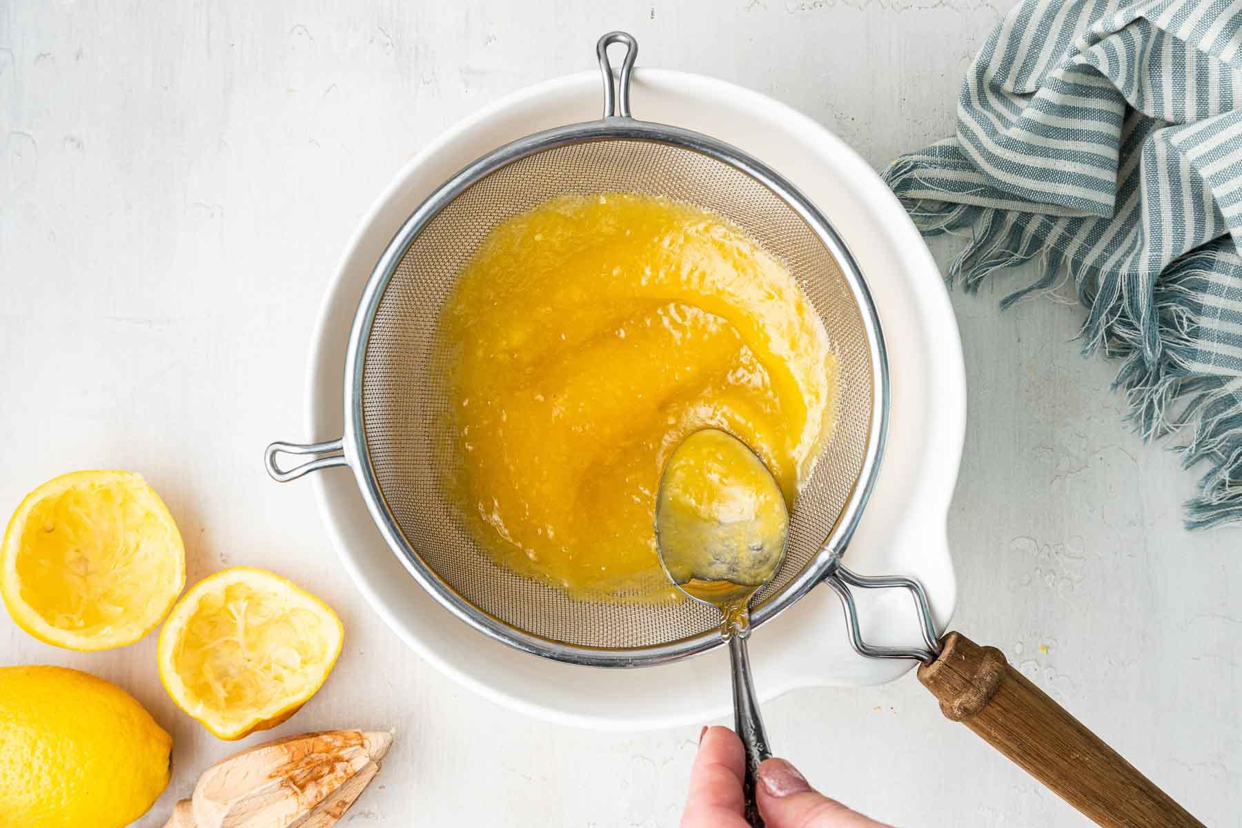 **Lemon Curd al Microonde in Piccola Quantità** 7 Cucchiaio che spinge la pastella gialla attraverso un colino sopra una ciotola bianca.