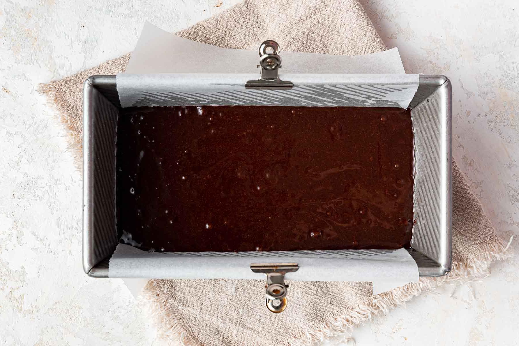 Impasto di brownies in piccola quantità versato in uno stampo di metallo da plumcake con carta forno e molletta di metallo sul lato.