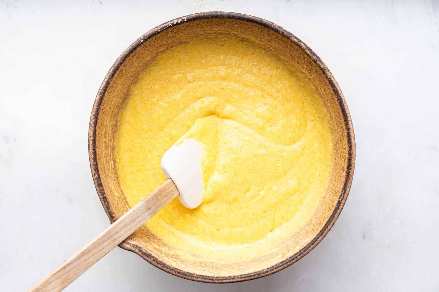 Ricetta Muffin al Mais 8 Impasto giallo in una ciotola marrone con una spatola bianca.