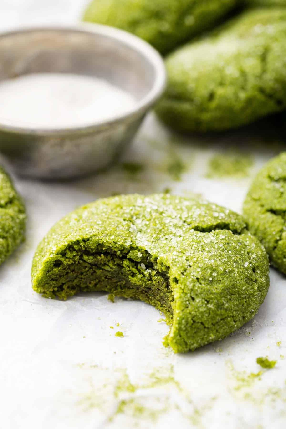 Biscotti al Tè Verde Matcha 9 Dolce verde con un morso mancante su un tavolo bianco accanto a una ciotola di zucchero.