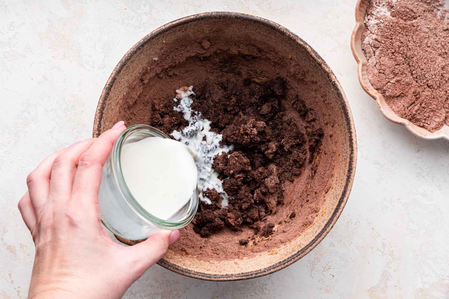 Hand pouring milk into brown dough in brown bowl.
