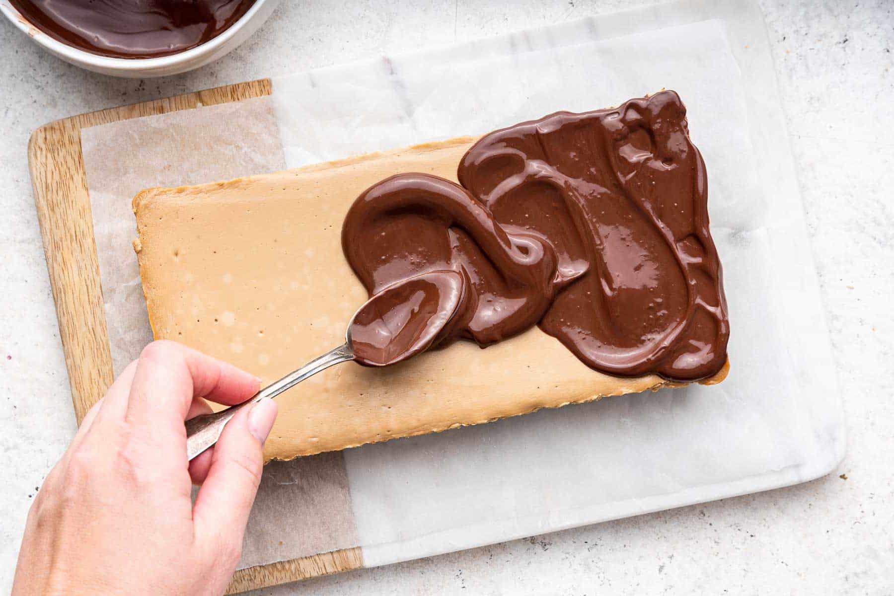Cheesecake al caffè 10 Mano che spalma la ganache al cioccolato su un dessert rettangolare marrone.