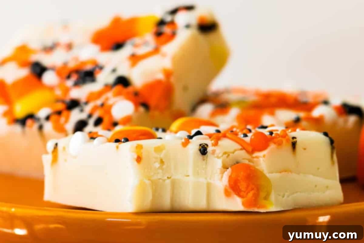 Haunted Fudge Delights 3 A close-up of a perfectly cut square of white chocolate Halloween fudge, with a bite taken out, revealing its creamy texture and embedded candy corn and sprinkles.