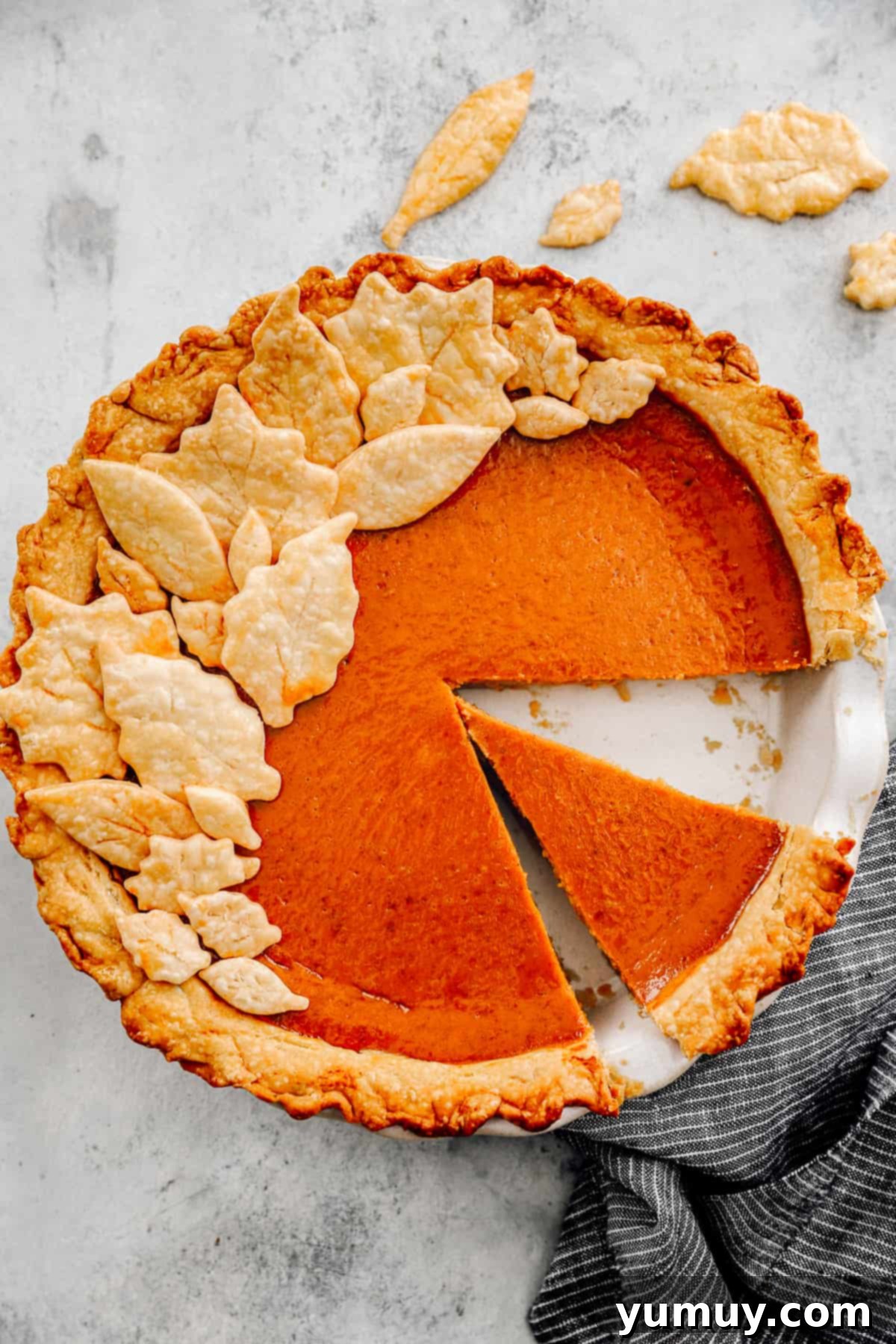 An overhead shot of a partially cut pumpkin pie, showcasing its smooth, creamy texture and a missing slice, adorned with decorative pie crust leaves.