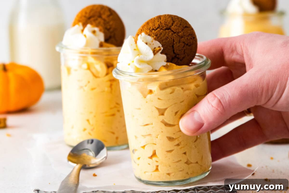 A hand gently reaching for a glass jar filled with perfectly chilled pumpkin mousse, topped with whipped cream and crumbled gingersnap cookies.
