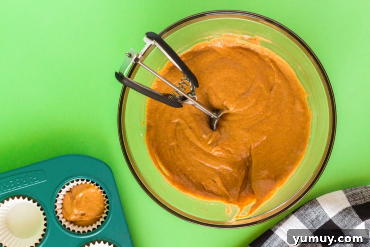 A glass bowl holds rich orange pumpkin cupcake batter, ready for scooping, with a metal cookie scoop resting beside it, highlighting the smooth texture and vibrant color.