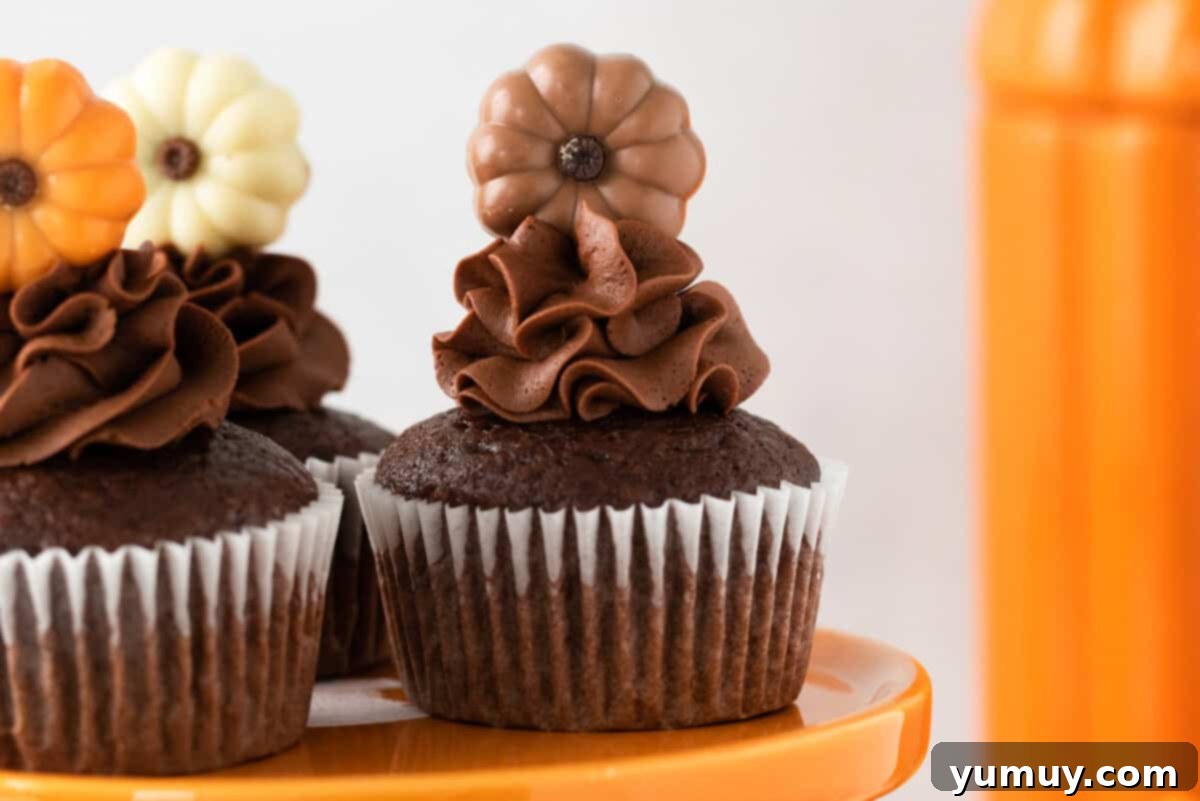 Close-up of three artfully frosted chocolate pumpkin cupcakes on an elegant orange cake stand, each adorned with a small, charming chocolate pumpkin decoration.