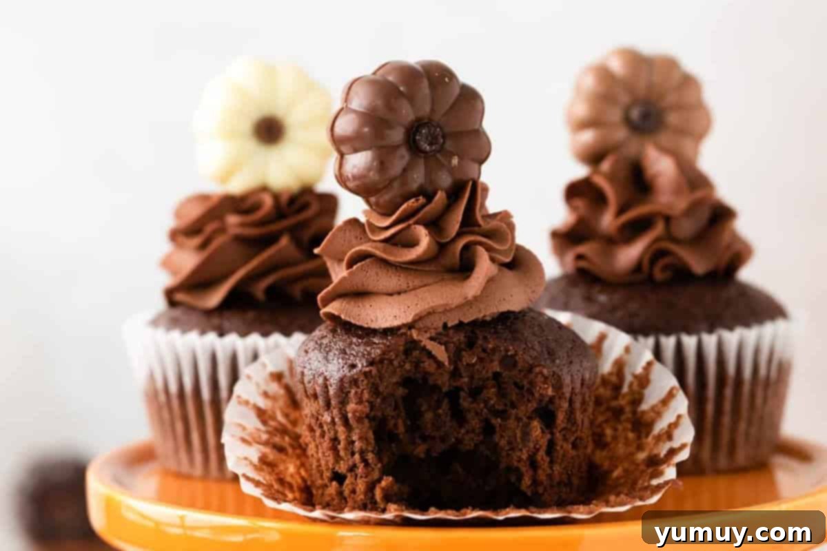 Close-up of a perfectly bitten chocolate pumpkin cupcake, revealing its moist, tender interior and luscious chocolate frosting, resting on an elegant orange cake stand alongside two other treats.