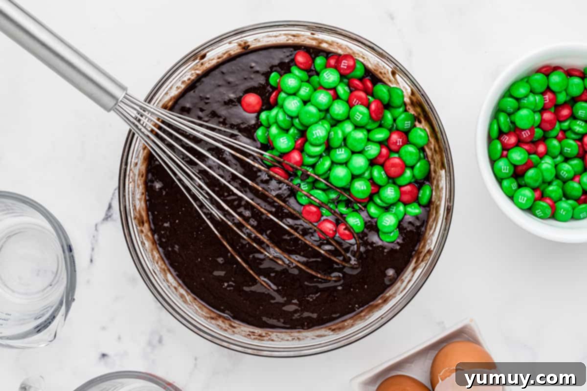 Brownie batter, generously dotted with vibrant Christmas M&M's, filling a white baking dish before going into the oven.