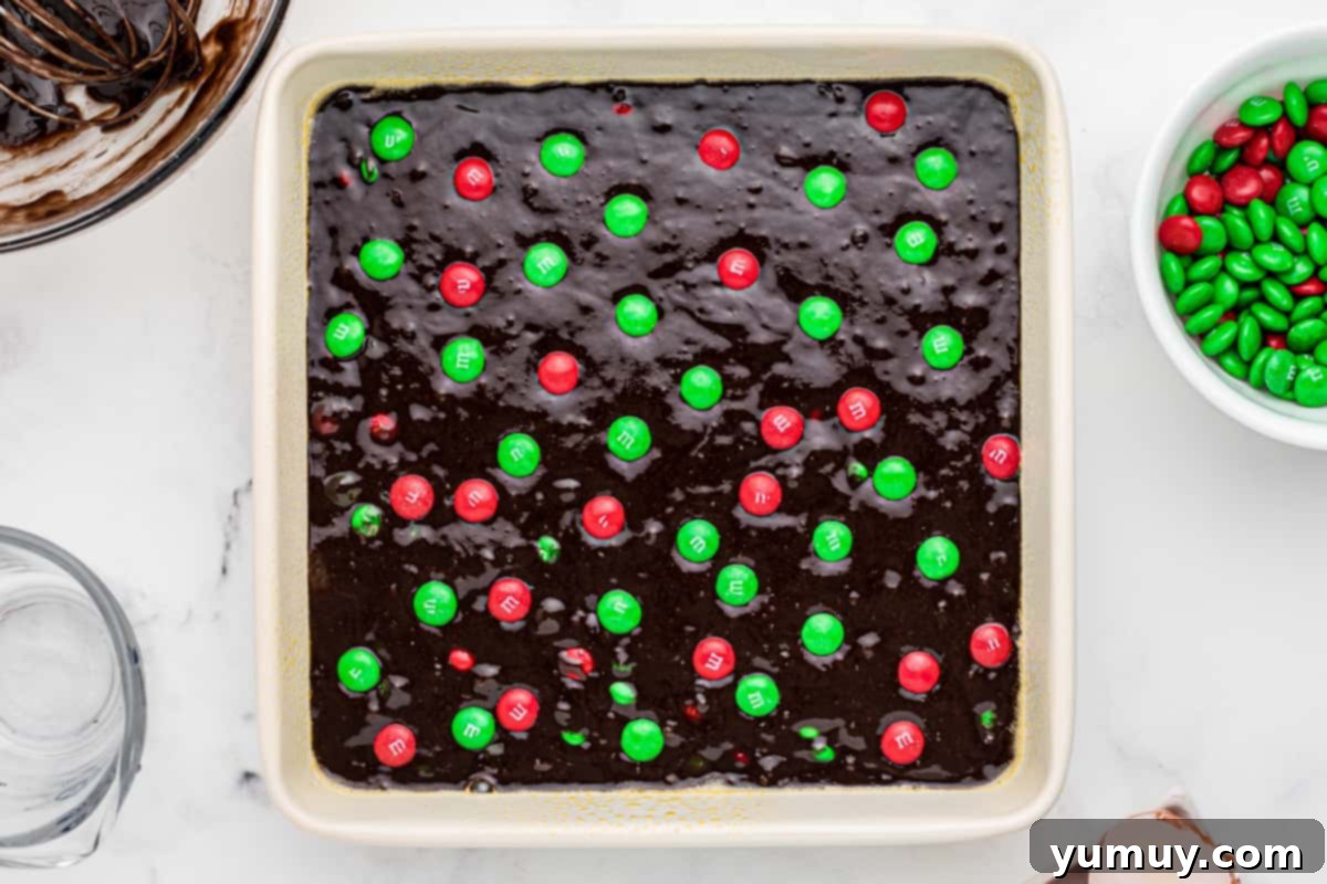 Unbaked brownie batter in a white baking dish, topped with festive Christmas M&M's, ready to be placed in the oven.