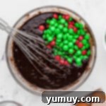 Rich brownie batter, speckled with colorful Christmas M&M's, filling a white baking dish.