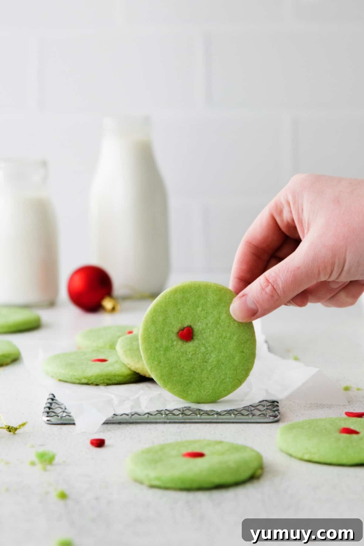 Hand holding up a green-colored Grinch sugar cookie with a red heart on it.