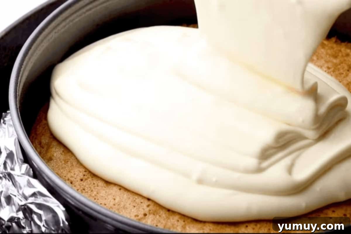 Pouring creamy cheesecake batter over the freshly baked carrot cake in a springform pan, preparing for the water bath.