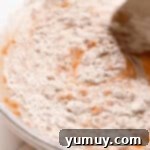 Stirring spiced flour into the mixture of carrots and eggs in a glass bowl, creating a rich carrot cake batter.