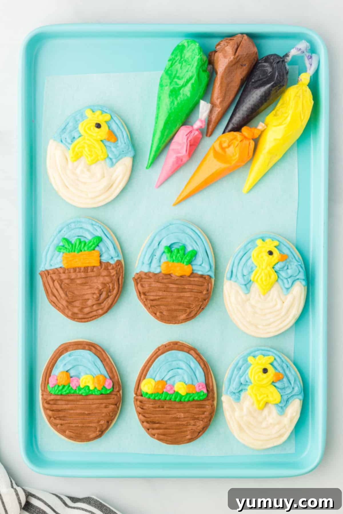 An overhead view of seven perfectly decorated Easter egg sugar cookies arranged on a blue baking sheet, alongside piping bags filled with colorful frosting.