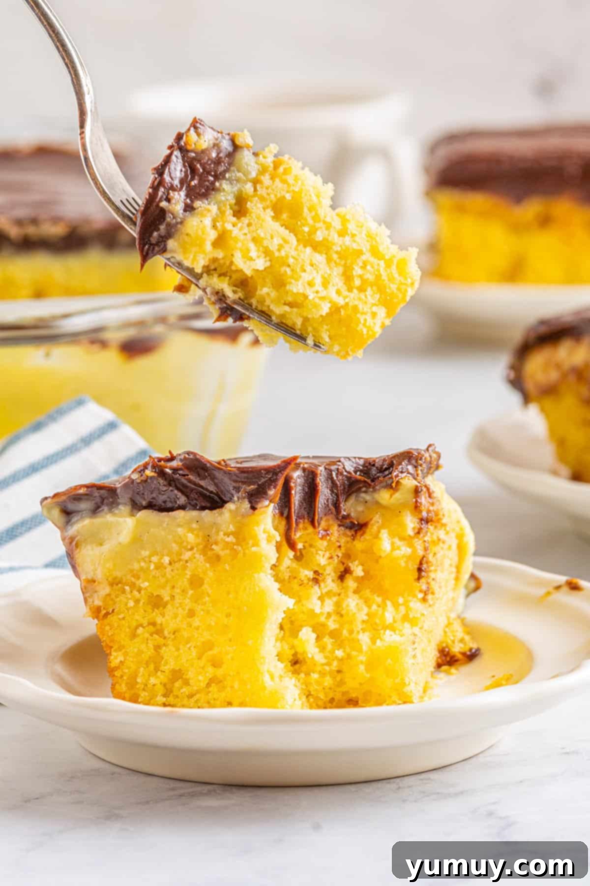 A forkful of Boston Cream Pie Poke Cake suspended above a slice on a white plate, showcasing its golden cake, creamy white filling, and dark chocolate topping.