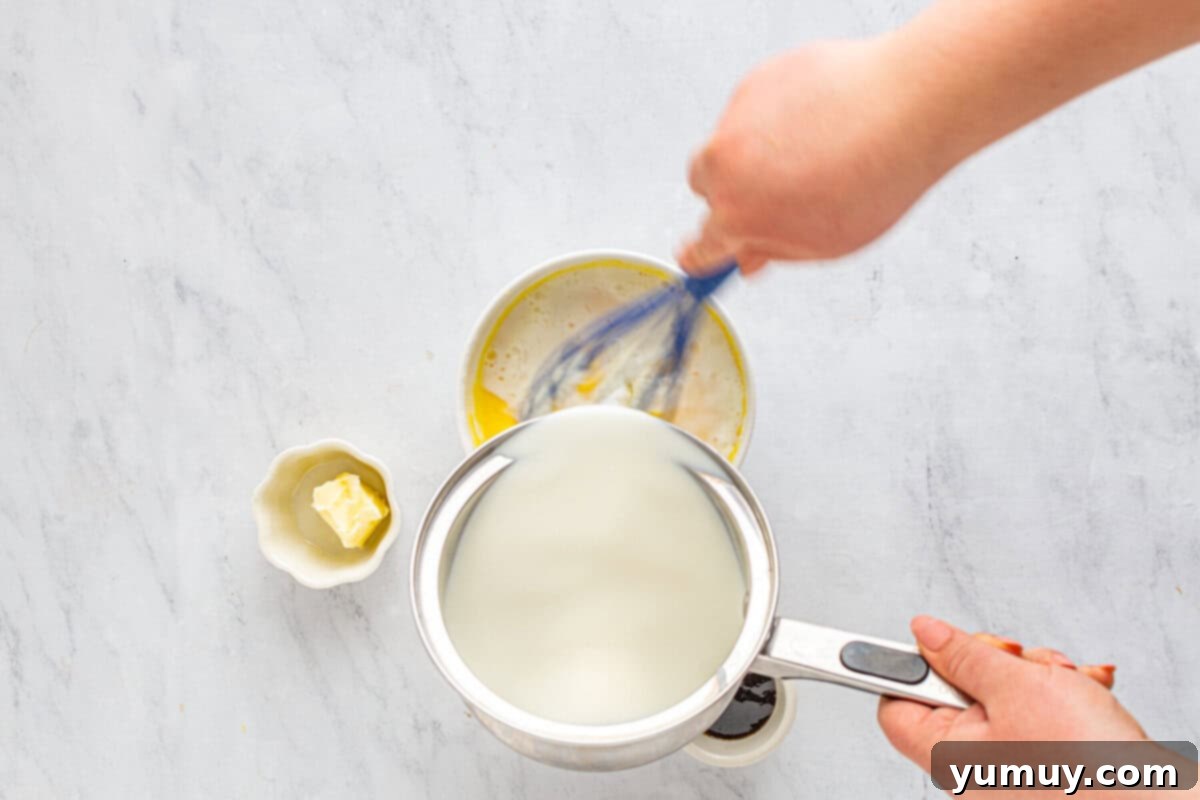 Hot, simmered milk being carefully poured in a slow stream into the egg yolk mixture, while simultaneously whisking to temper the eggs for the pastry cream.