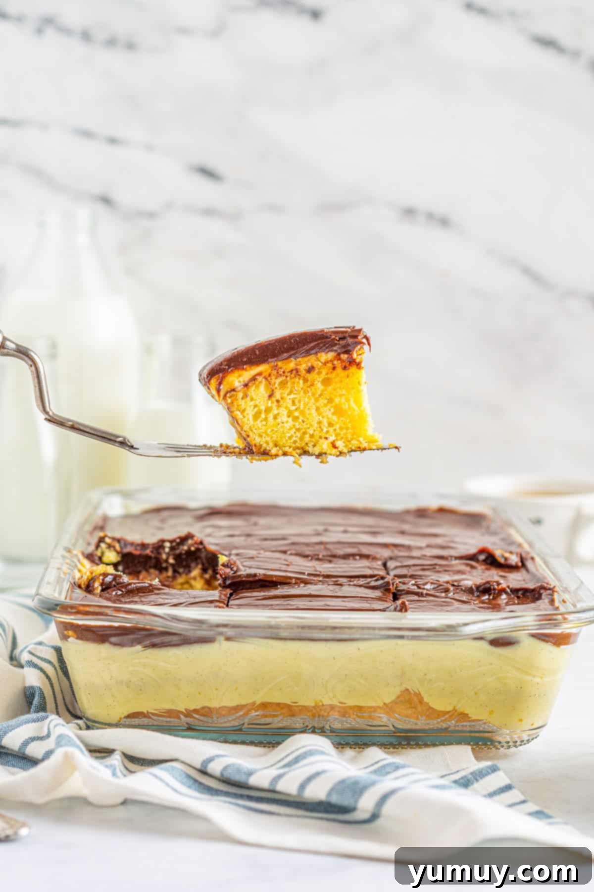 A perfectly sliced portion of Boston Cream Pie Poke Cake being lifted on a cake server, showing the distinct cake, cream, and ganache layers against the backdrop of the whole cake.