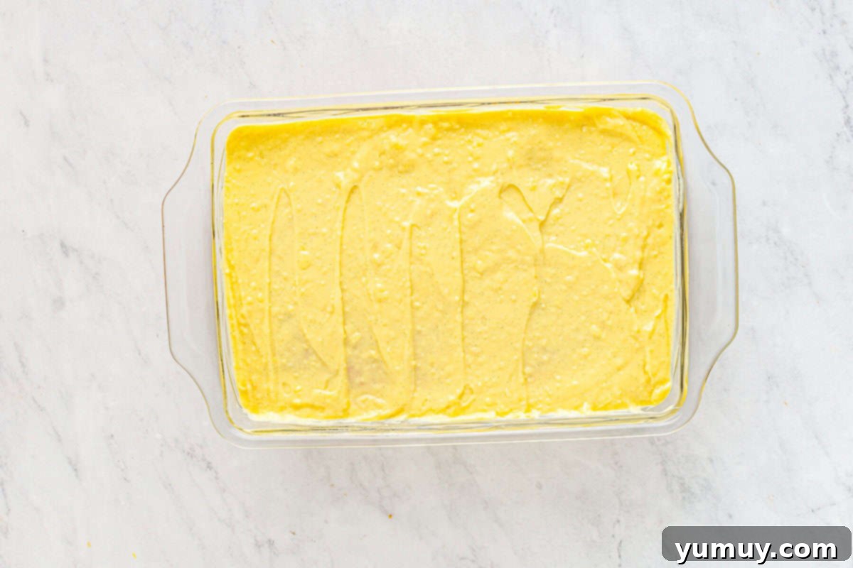 Thick pastry cream generously layered over the poked yellow cake in a glass baking dish, smoothly spread and ready for its essential chilling period.