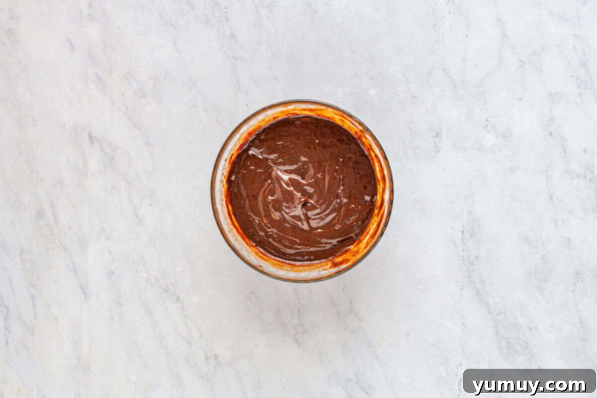 Freshly prepared, smooth, and glossy chocolate ganache in a clear glass bowl, ready to be poured over the chilled cake.