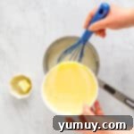 The tempered egg mixture being poured back into the saucepan with the remaining hot milk, to be whisked together with a blue whisk.
