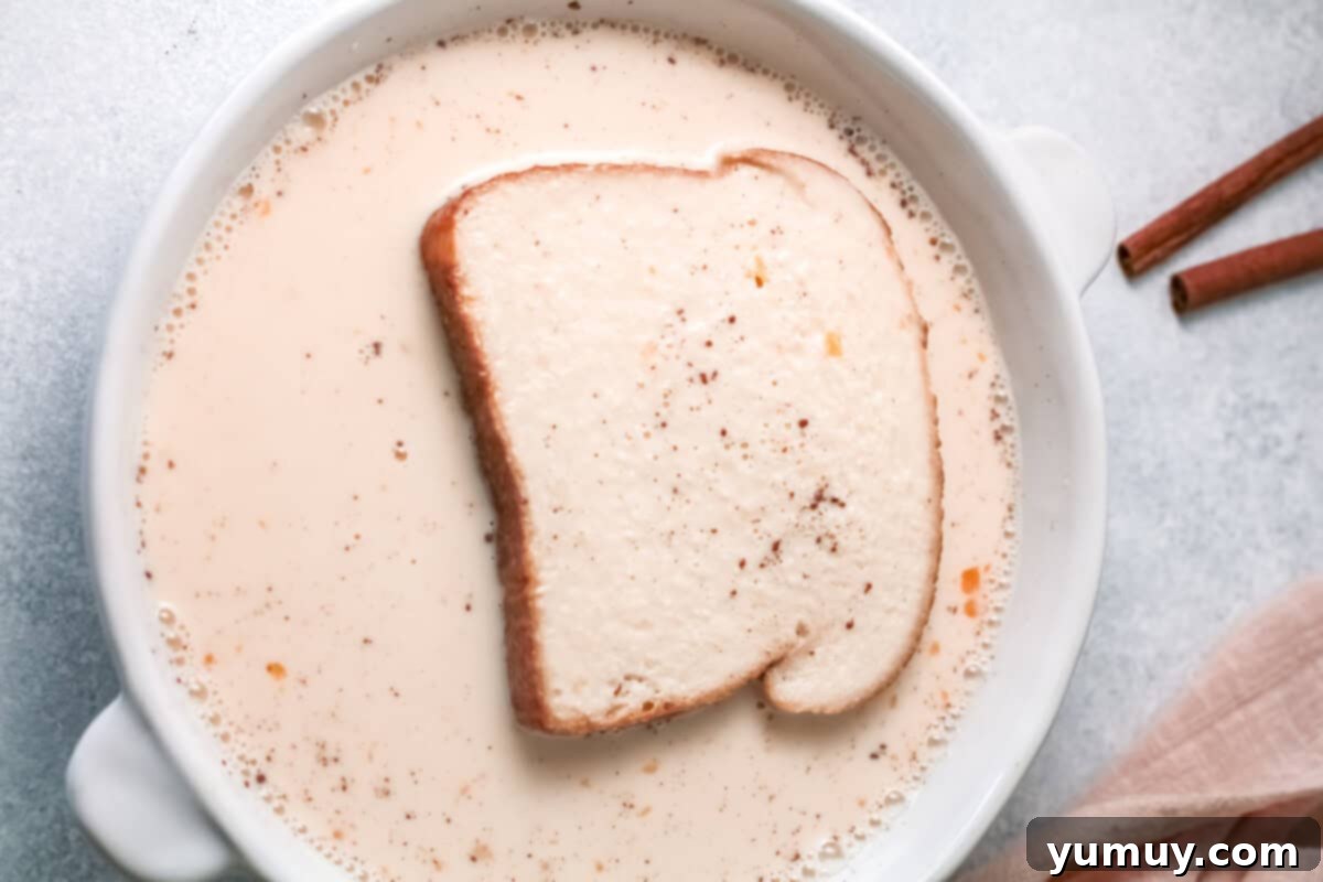 a slice of brioche bread being dipped in the rich egg mixture in a white bowl.