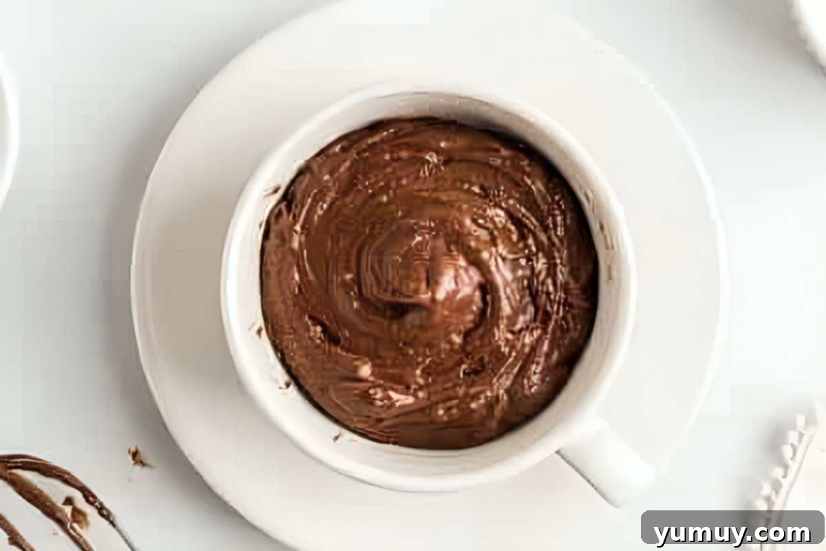 Smooth chocolate cake batter resting in a white coffee mug, ready for topping and microwaving.