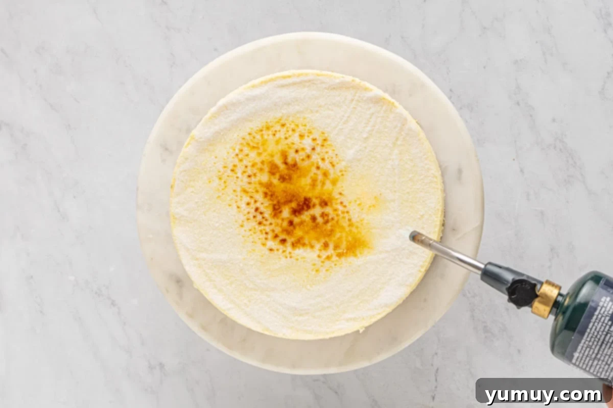 a kitchen torch caramelizing sugar on top of a cheesecake on a white cake stand.