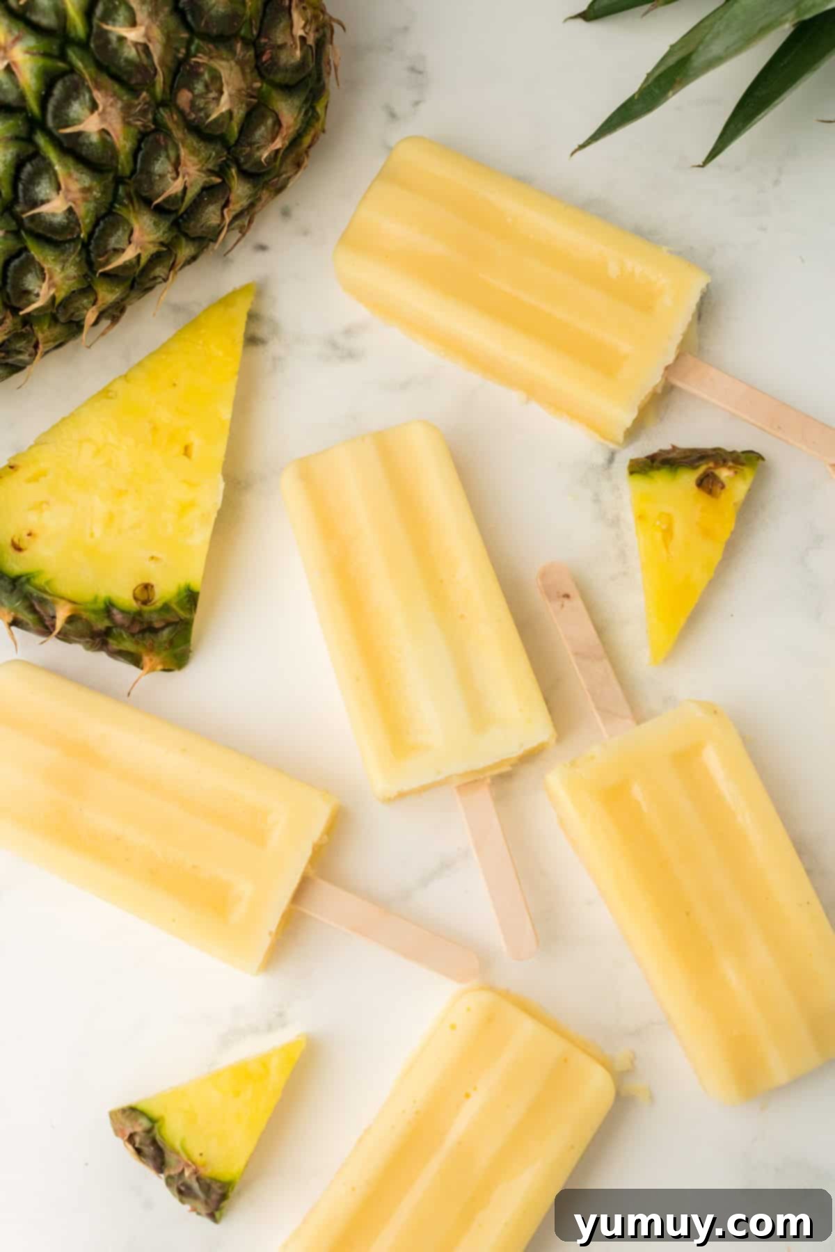 Creamy Dole Whip popsicles laid on a countertop, surrounded by fresh pineapple slices, highlighting their tropical appeal.