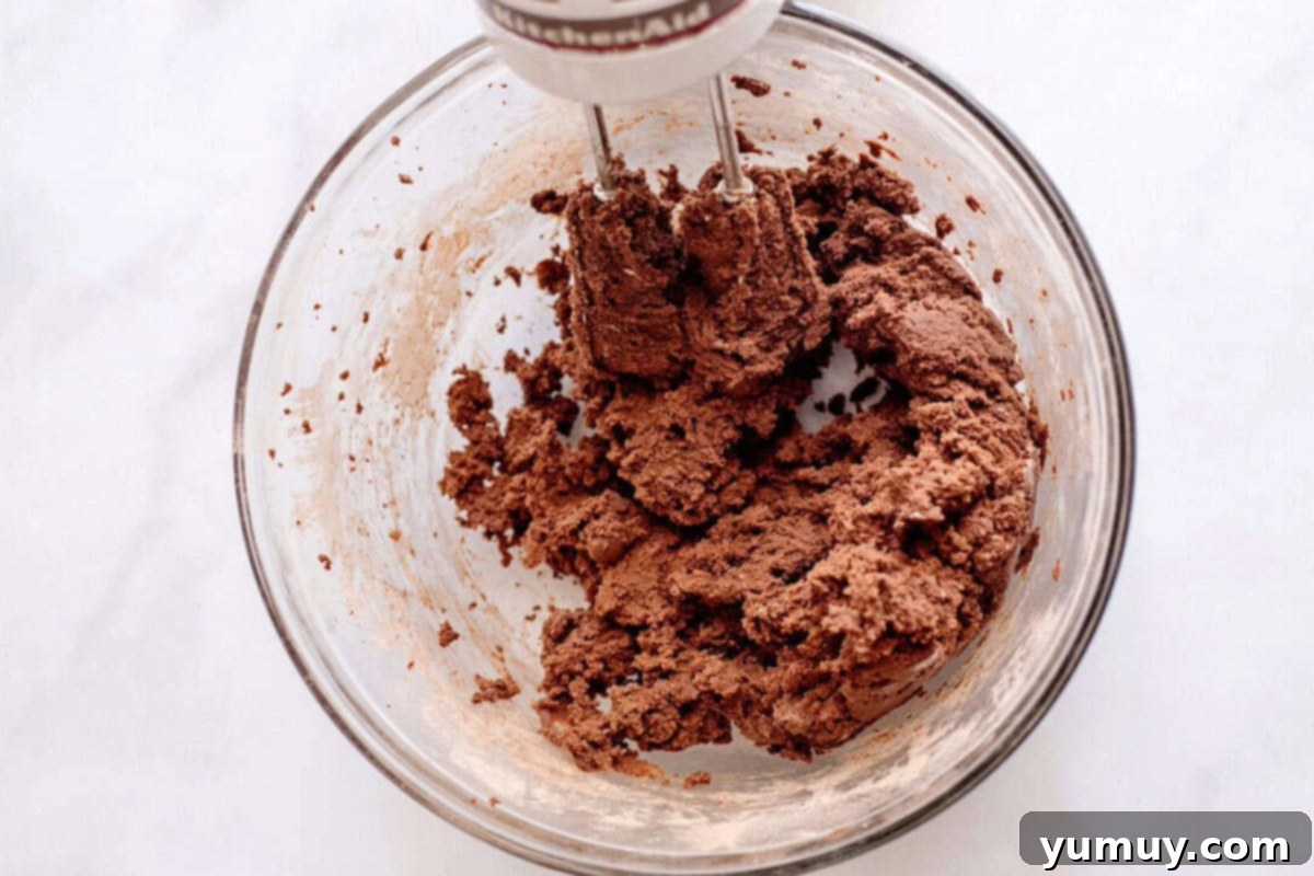 Gooey Chocolate Marshmallow Cookies 17 Mixing chocolate cookie dough with a hand mixer, demonstrating the careful process of incorporating dry into wet ingredients.