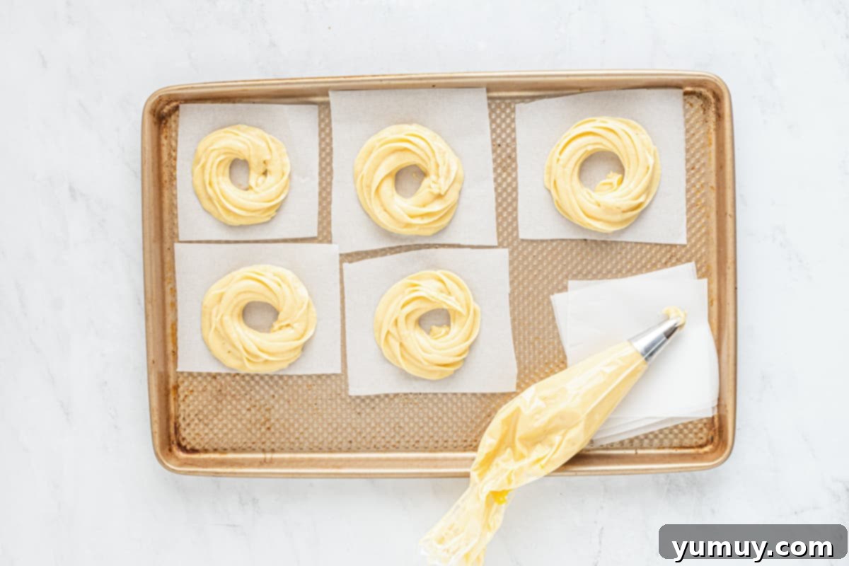 Piping cruller batter into uniform ring shapes onto parchment paper squares on a baking tray.