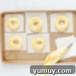 Piping cruller dough into ring shapes on parchment paper on a baking tray.