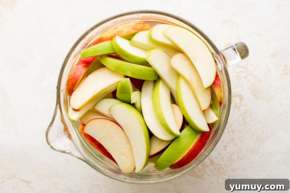 Crisp apple slices resting in a large glass measuring bowl, completely covered with cold water to prevent browning and maintain freshness.