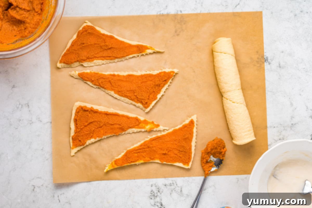Triangles of crescent roll dough are laid flat on parchment paper, with a spoonful of orange pumpkin pie filling on each.