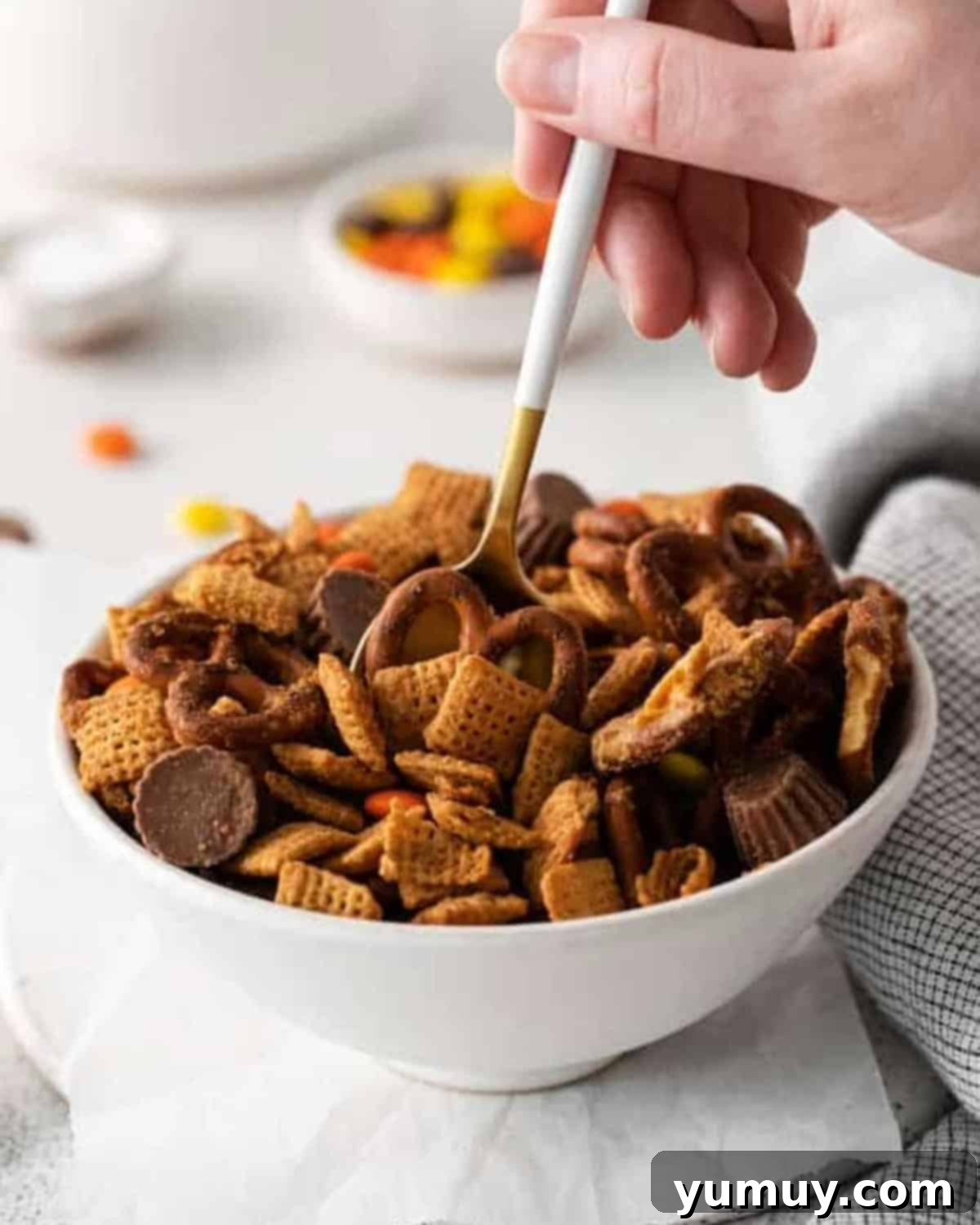 A spoon scooping a generous portion of chocolate peanut butter Chex mix from a pristine white bowl, highlighting its abundant mix-ins.