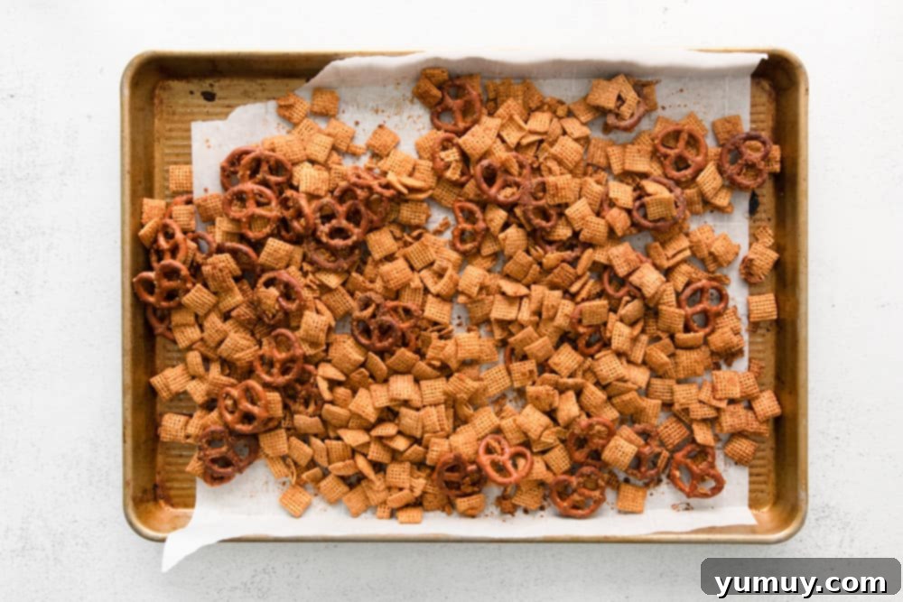 Peanut butter Chex and pretzels spread in a single layer on a parchment-lined baking sheet, ready for the oven.
