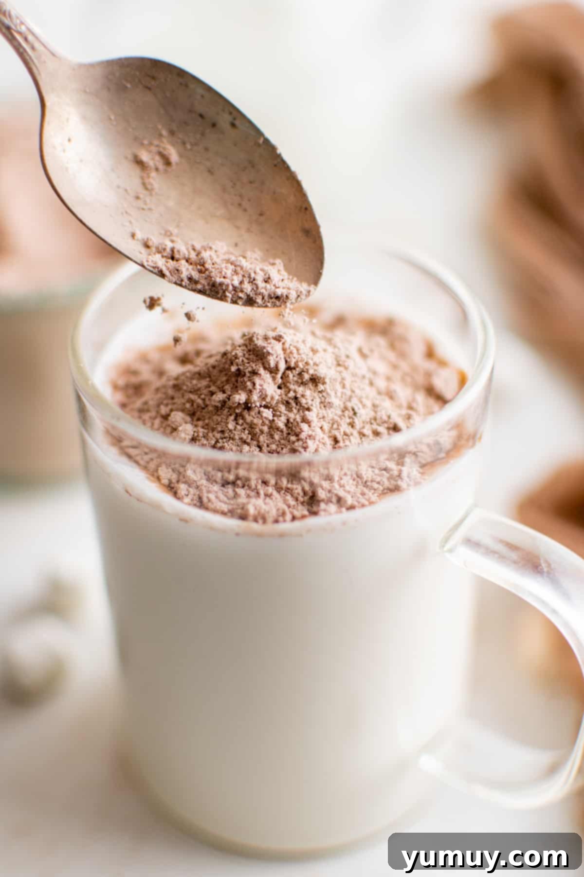 A spoon pouring homemade hot chocolate mix over a glass of milk, ready to be stirred into a rich, creamy beverage.