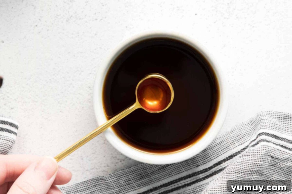 A teaspoonful of homemade vanilla extract held over a bowl of extract, showcasing its dark, rich color.