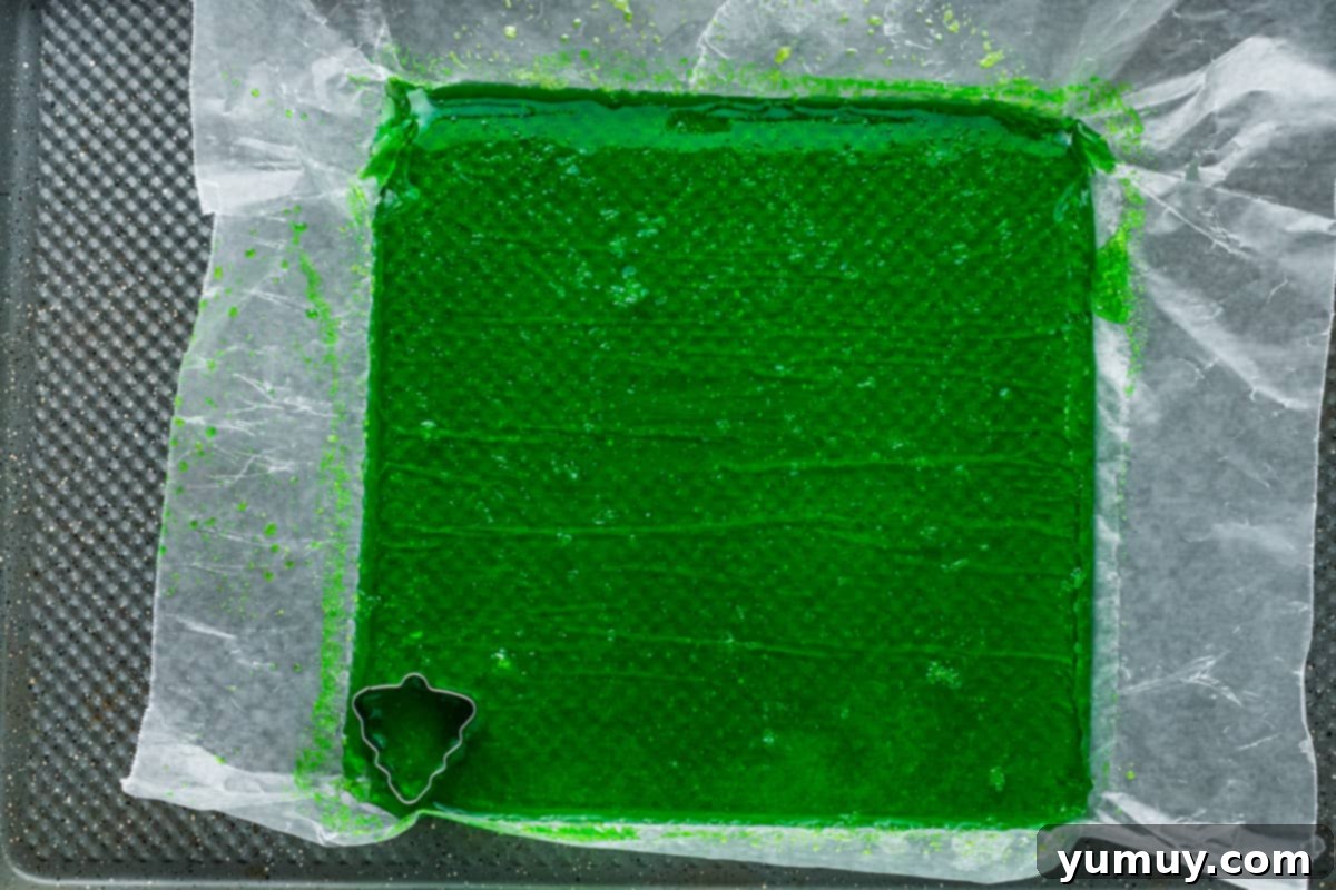 A sheet of green gumdrop candy on parchment paper, with a Christmas tree cookie cutter poised for cutting a festive shape.